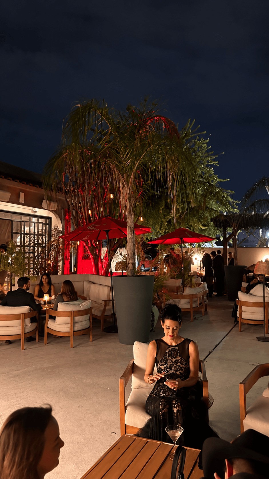 Guests at a candlelit tropical lounge at night, under palm trees and red umbrellas